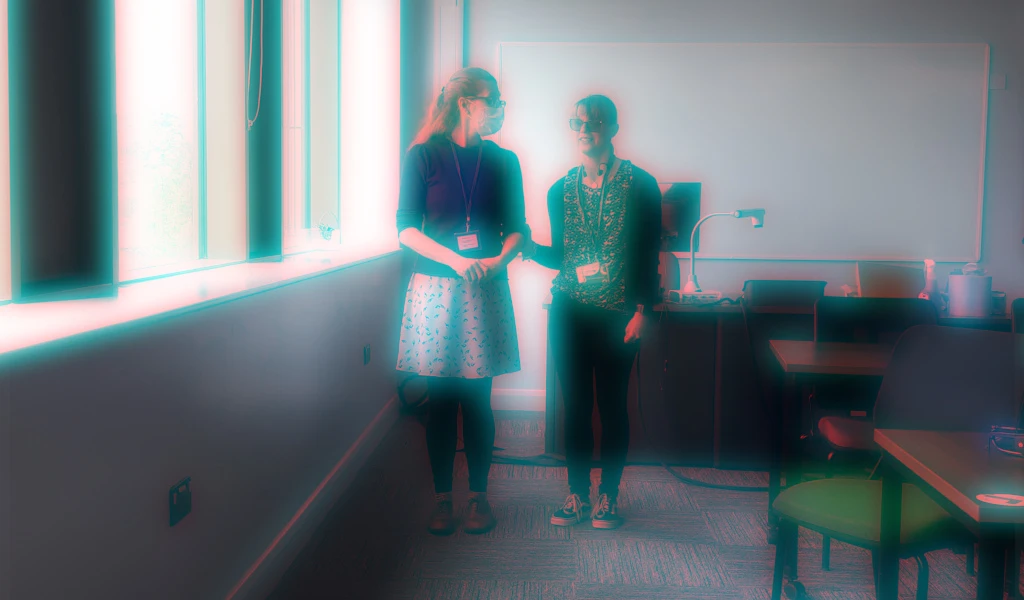 Vicky and Jade standing in a room facing us with a whiteboard behind them, windows on their right, and tables on the left. The image is blurred.