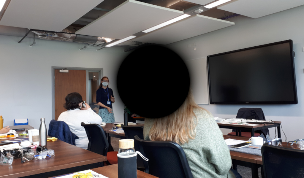 Vicky standing in front of people sitting at desks. The image in the centre is blackened with a circular shape to simulate macular degeneration.