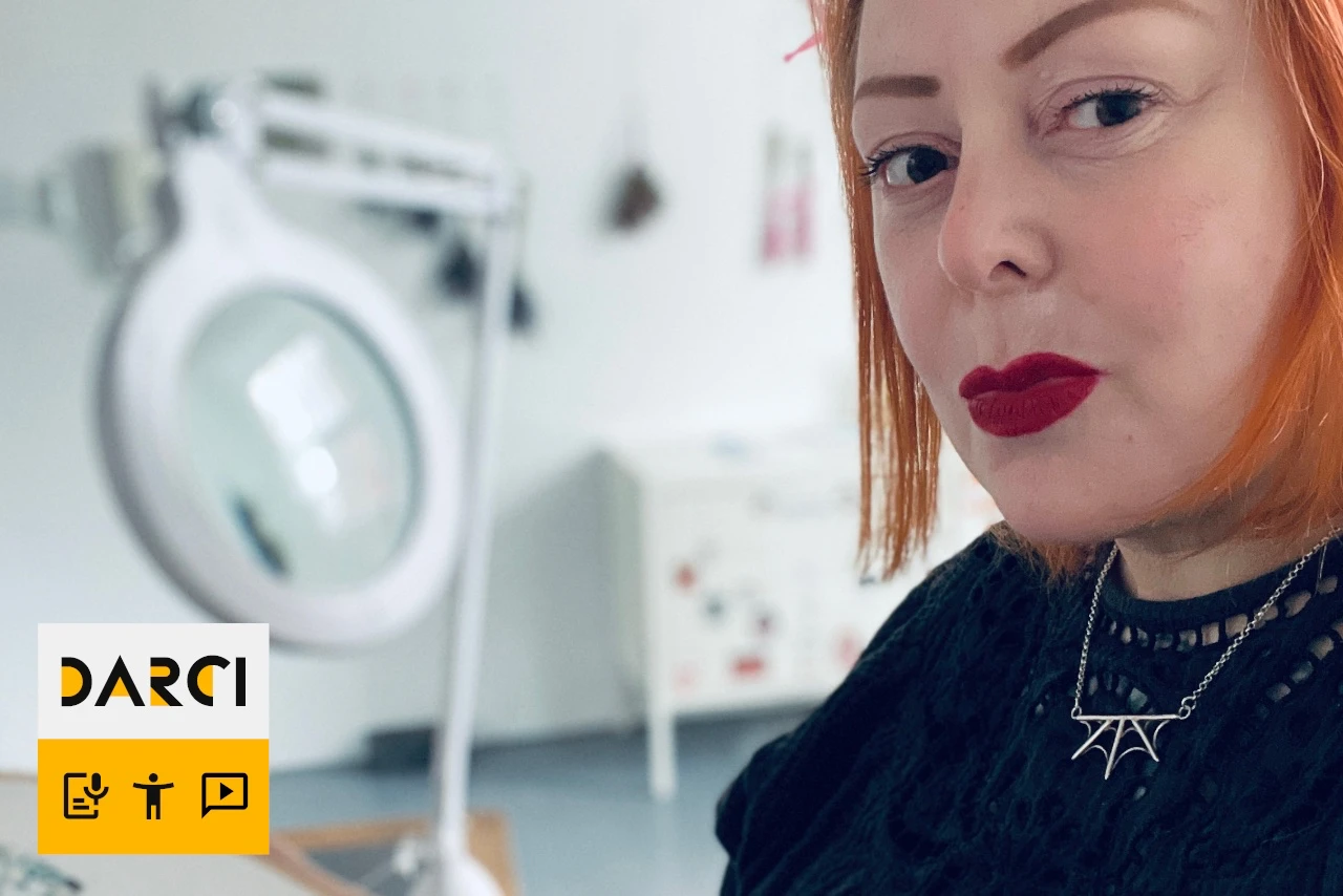 The artist, Michelle Duxbury, in her studio. Wearing a black dress she is sat to the right of the image holding a large embroidery hoop with green thread attached. She is a white woman with ginger hair and bright red lipstick. On a desk behind her is a large white microscope. The room is white with grey floors and has a small white cabinet against the back wall.