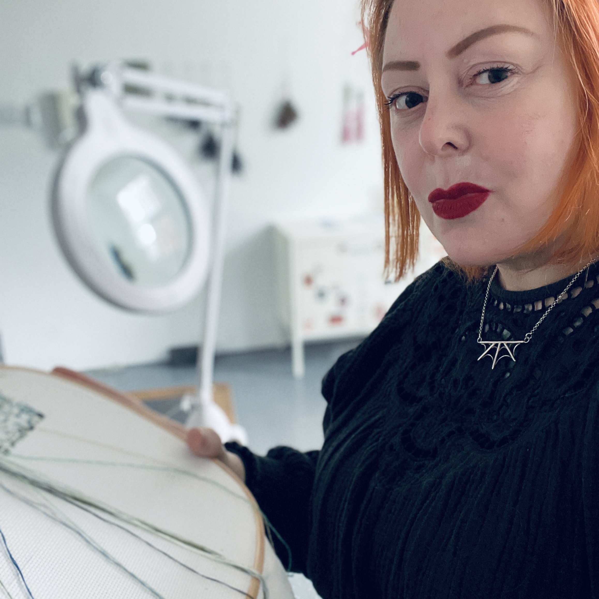 The artist, Michelle Duxbury, in her studio. Wearing a black dress she is sat to the right of the image holding a large embroidery hoop with green thread attached. She is a white woman with ginger hair and bright red lipstick. On a desk behind her is a large white microscope. The room is white with grey floors and has a small white cabinet against the back wall.