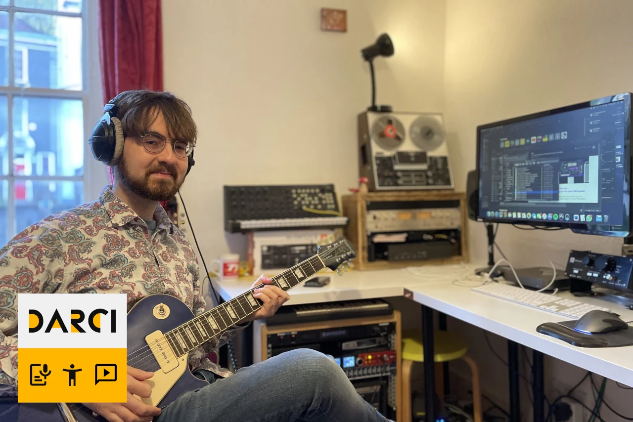 Photo of Jay sitting and holding a guitar. He has a computer on a desk next to him.