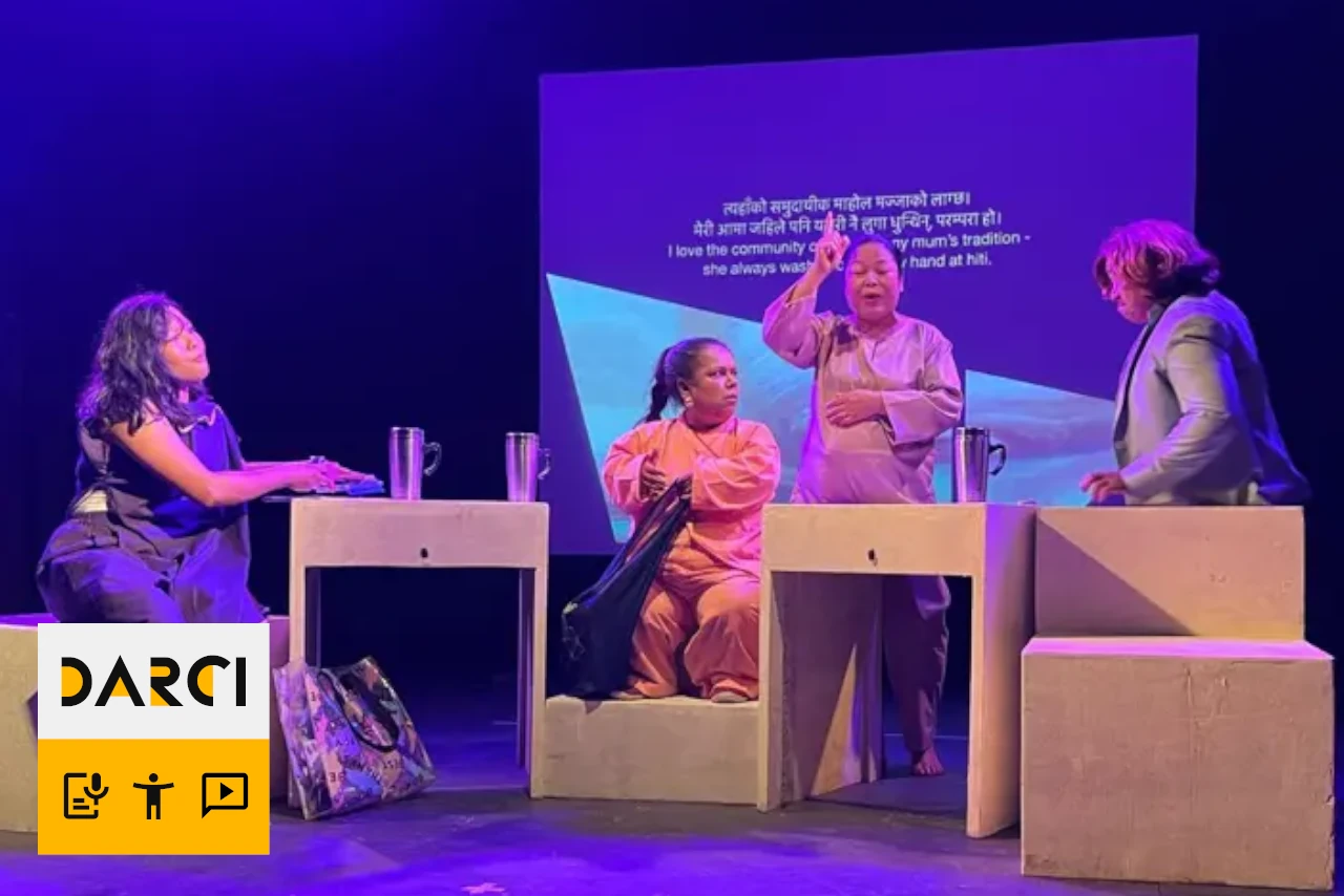 Four women on stage perform a scene, sitting and standing around rectangular blocks, with a translated text projected on a screen behind them.