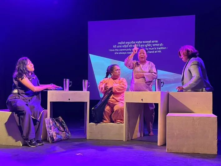 Four women on stage perform a scene, sitting and standing around rectangular blocks, with a translated text projected on a screen behind them.