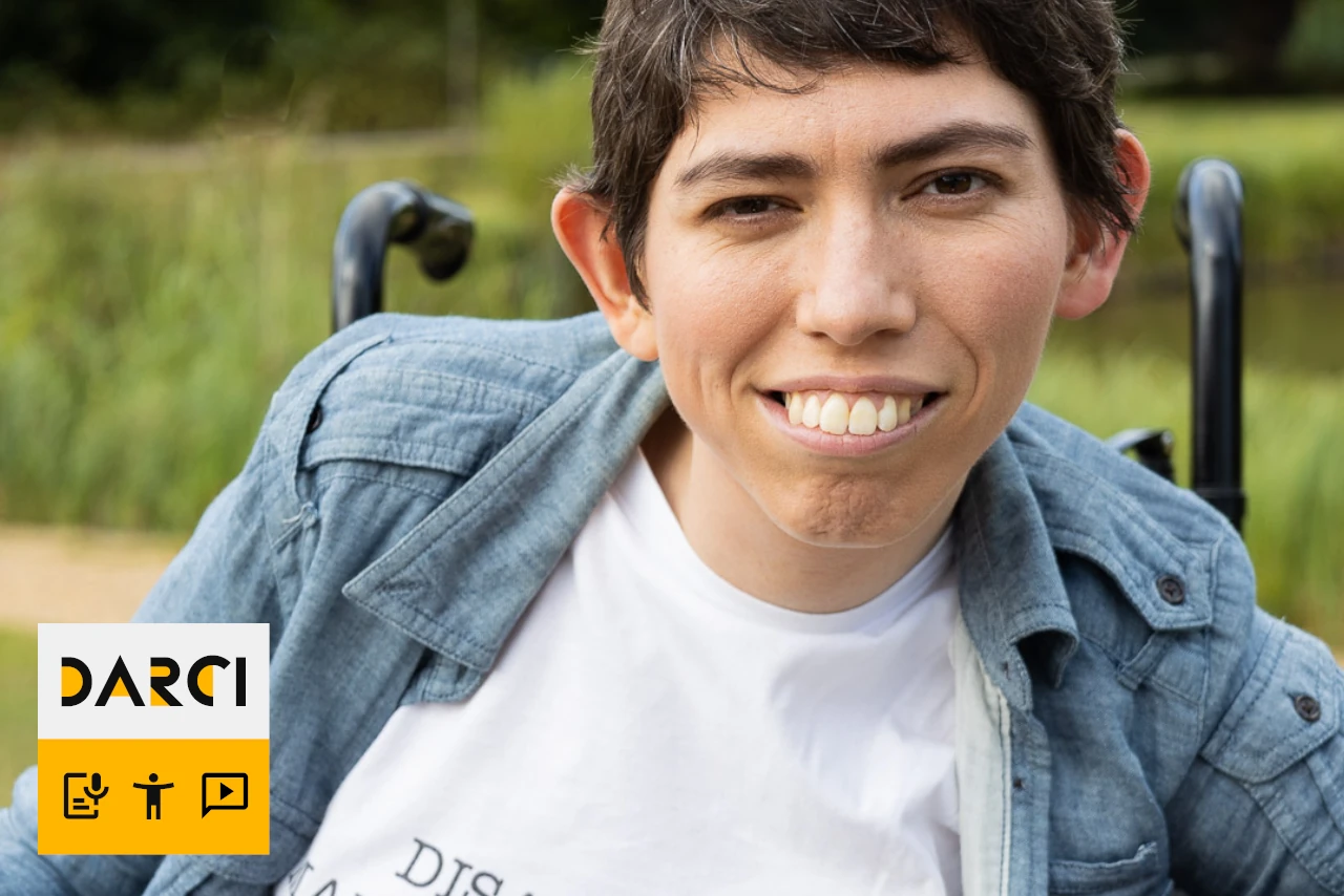 A person in a wheelchair smiles outdoors, wearing a shirt that reads DISABLED MAIN CHARACTER.