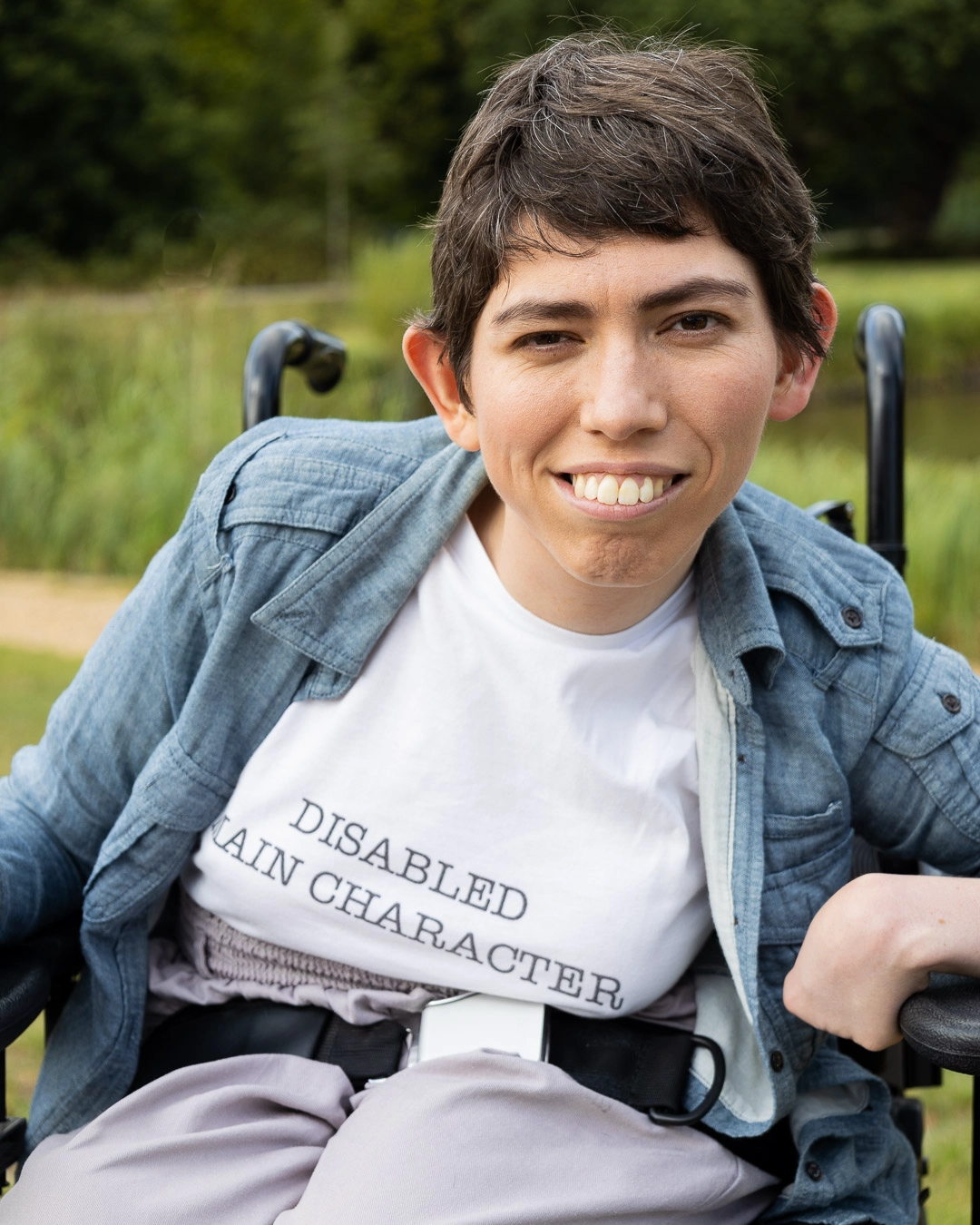 A person in a wheelchair smiles outdoors, wearing a shirt that reads DISABLED MAIN CHARACTER.