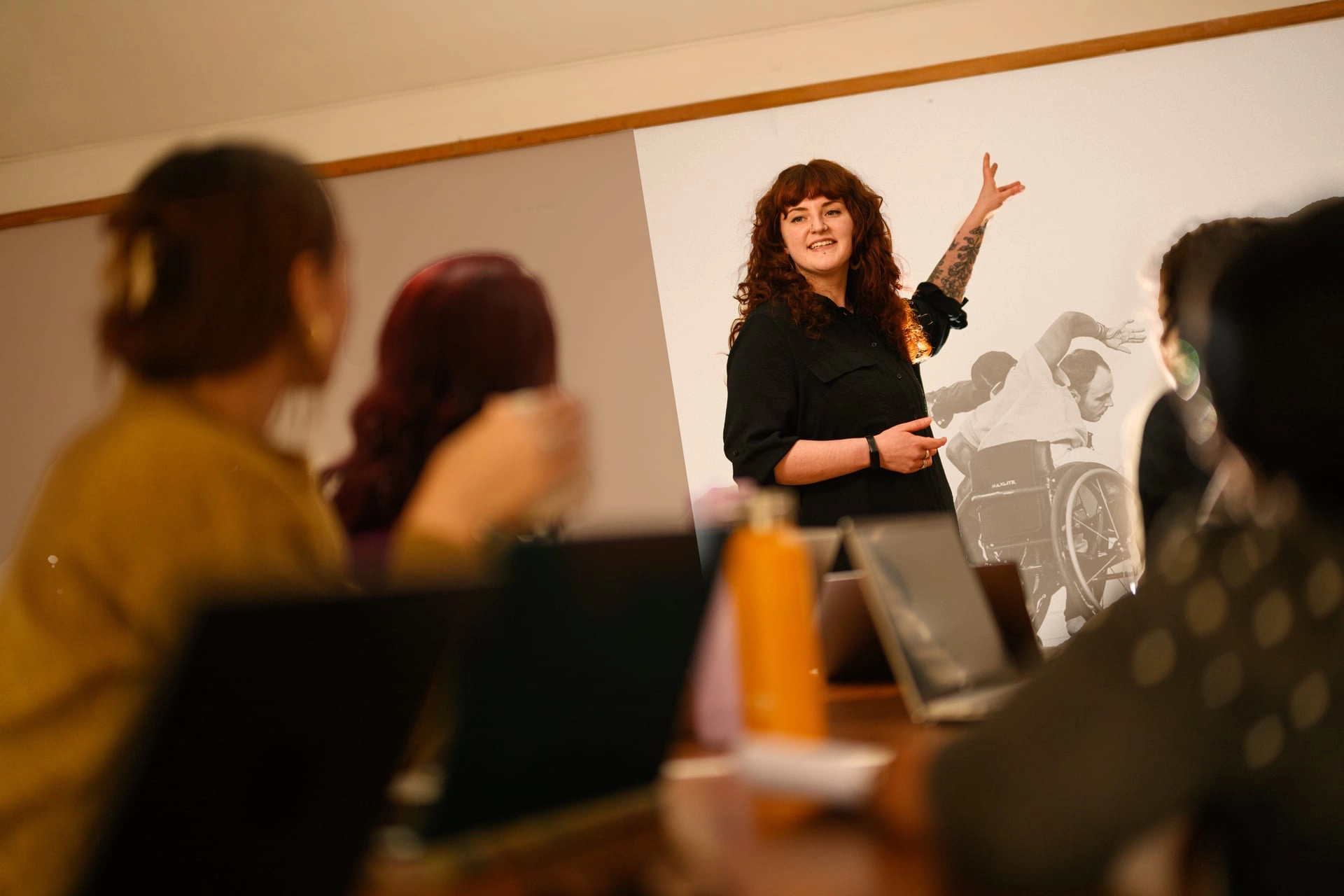 Lily stands and gestures toward a projected image on a screen while others sit at a table with laptops, watching her presentation.