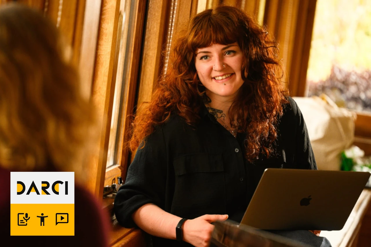 Lily with curly red hair smiles while holding a laptop, sitting by a window. The DARCI logo is shown in the lower-left corner of the image.