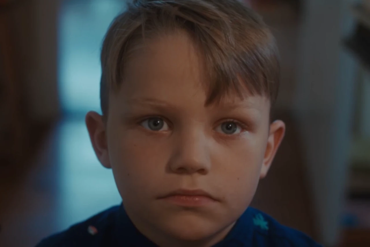 ‘A young boy with short, light-brown hair and blue eyes, gazes intently at the camera. The background is softly blurred, adding a serious tone.’