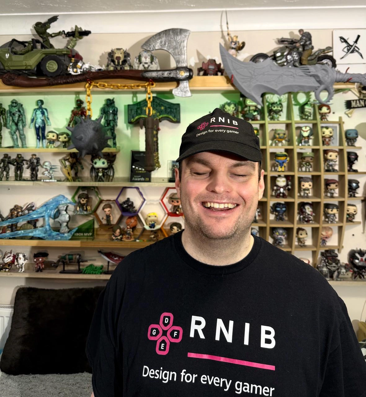 SK wearing an RNIB t-shirt and hat stands in front of shelves filled with action figures and gaming collectibles.