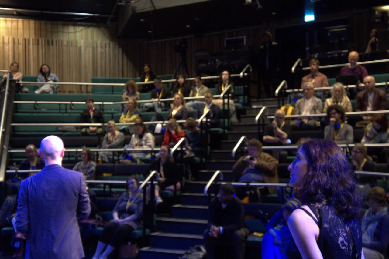 Mariana and Gavin stand before an audience seated in tiered rows inside a modern lecture hall or theater, with most attendees looking toward the stage.