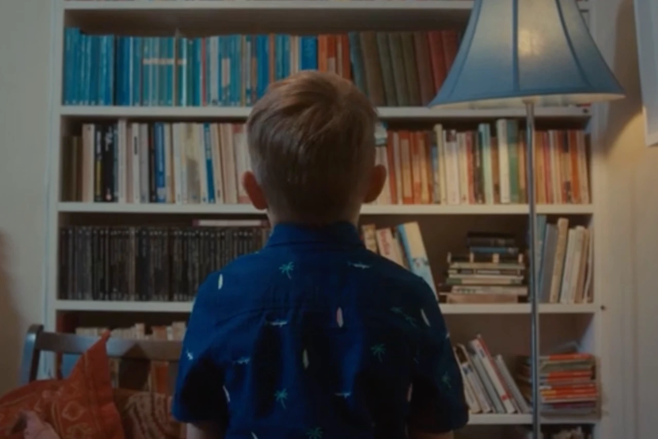 A young boy stands in front of a tall bookcase filled with colorful books.